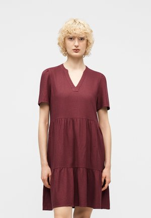 Blondynka w bordo sukience z krótkim rękawem, sięgającej kolan, z dekoltem w serek i rozkloszowanym, marszczonym dołem, stojąca na jednolitym tle.