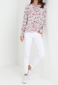 Blusenshirt mit langen Ärmeln in floralem Design auf weißem Grund und rosa Akzenten, kombiniert mit schmal geschnittenen weißen Jeans und weißen Sneakers. Strukturierter, lässiger Stil.