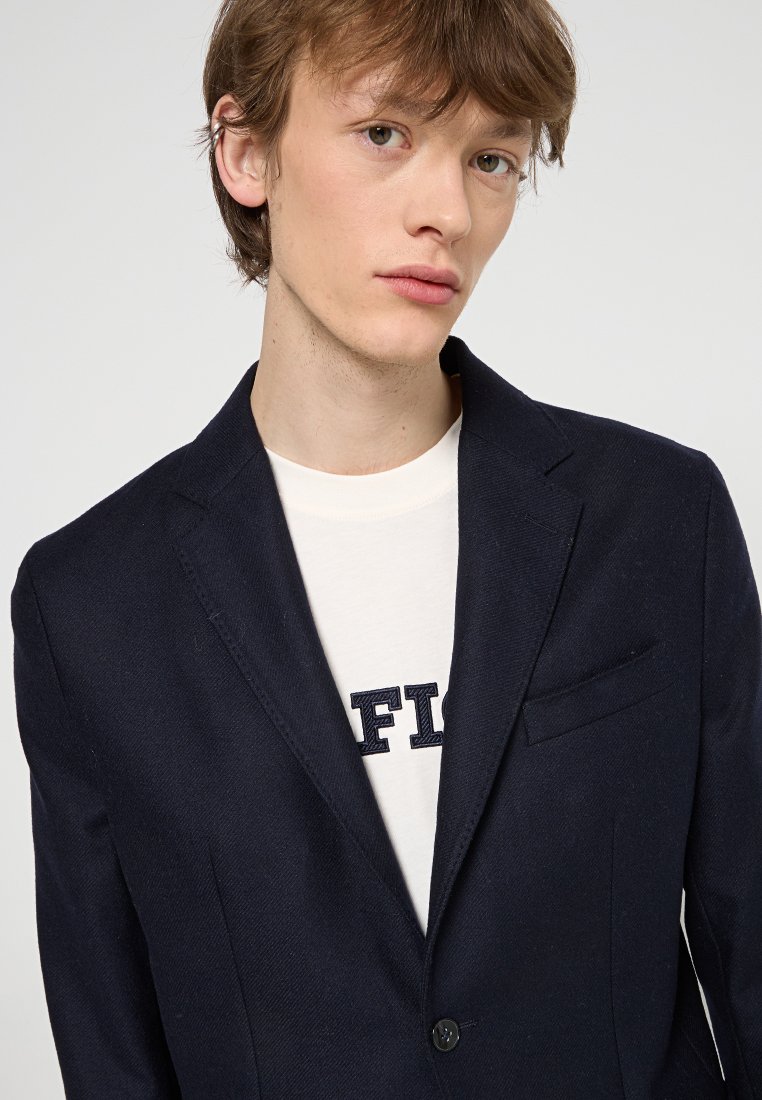 Granatowa marynarka o dopasowanym kroju, z zapięciem na pojedynczy guzik i klapami w kształcie lenona. Noszona na kremowej koszuli z niebieskim napisem.