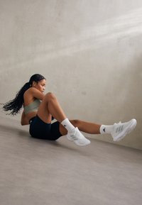 Calzature sportive bianche con dettagli grigi, abbinate a calzini bianchi con logo nero. Il modello indossa un reggiseno sportivo verde chiaro e pantaloni corti neri.