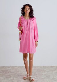 Rosa bomullsklänning med långa ärmar och en nyckelhålsringning. Matchad med en flätad gul handväska och ljusbeiga sandaler. Slät textur.