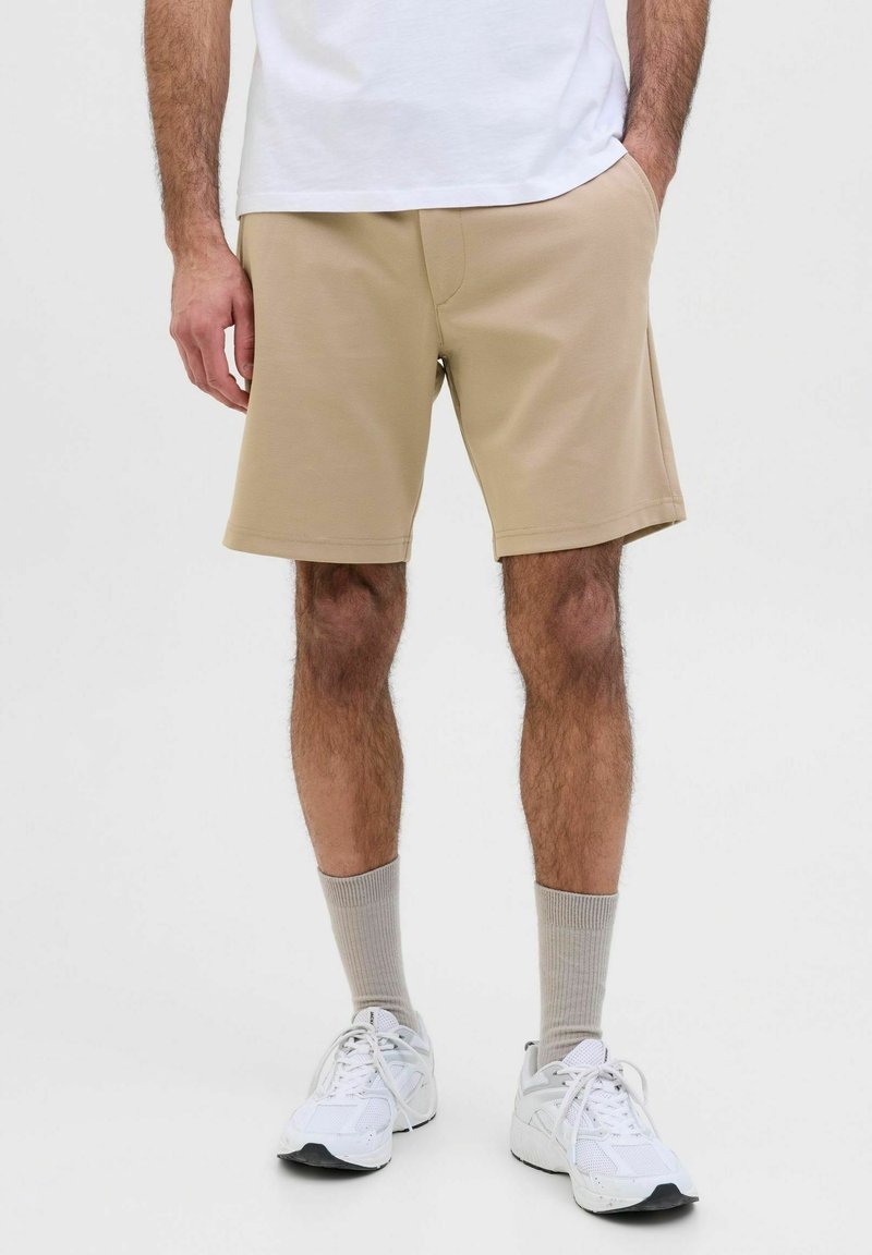 Homme portant un short beige, des baskets blanches, des chaussettes grises et un t-shirt blanc, debout les mains dans les poches.