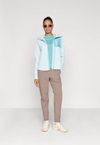 Ανοιχτό μπλε τζάκετ με φερμουάρ και ροζ λεπτομέρειες πάνω από τούρκιζ τοπ, συνδυασμένο με μπεζ παντελόνι και μπεζ αθλητικά παπούτσια. Το μοντέλο φοράει γυαλιά ηλίου.