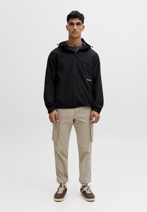 Ung mand stående iført en sort hættetrøje med lynlås, beige cargobukser og brune sneakers med hvide snørebånd på en ensfarvet baggrund.