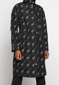 Czarny długi płaszcz z białym powtarzającym się wzorem "Hugo", zapięcie na zatrzaski i ściągnięta talia, noszony na czarnych spodniach.