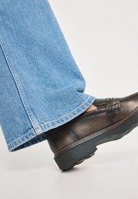 Zapato de cuero marrón texturizado con un tacón bajo, combinado con unos vaqueros de denim azul claro anchos que presentan un dobladillo deshilachado.