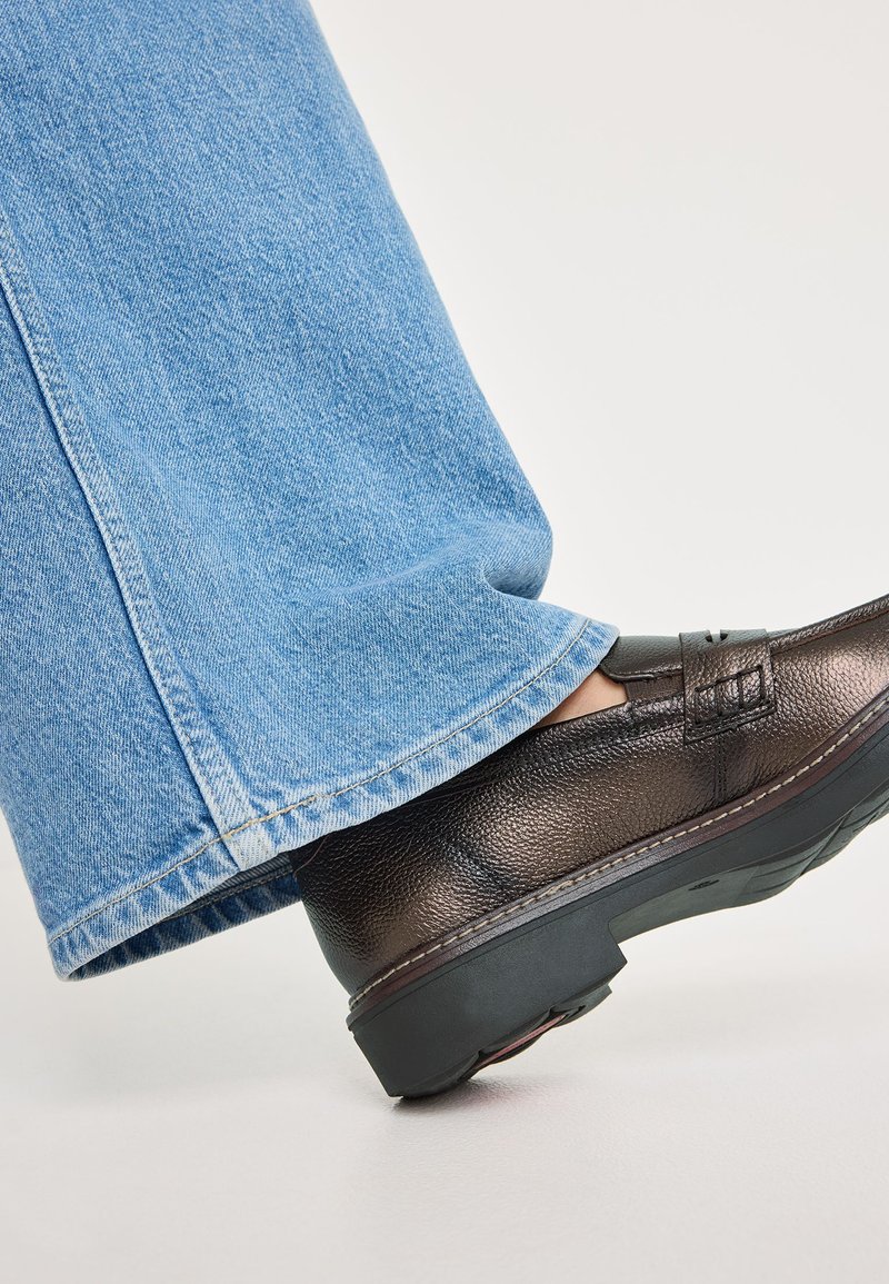 Zapato de cuero marrón texturizado con un tacón bajo, combinado con unos vaqueros de denim azul claro anchos que presentan un dobladillo deshilachado.