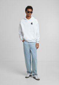 Haut à capuche blanc avec logo noir, poche kangourou, coupe décontractée. Jean droit bleu clair. Baskets grises et blanches avec accents noirs.