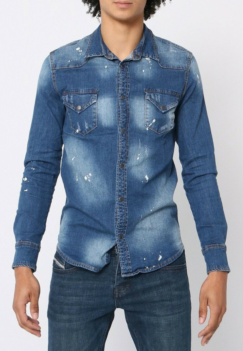 Chemise en denim de finition bleue usée avec des manches longues, un col boutonné à l'avant, deux poches sur la poitrine et des détails de éclaboussures de peinture.