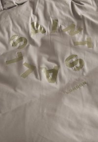 Ljust grått tyg med rynkig textur som har djärva, metalliska guldbokstäver som stavar "SAINTS" och "LONDON" längst ner.