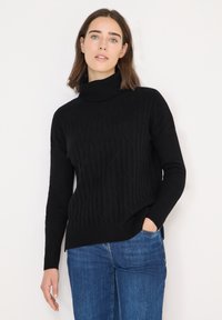 Pull à col roulé noir en maille côtelée, avec une coupe décontractée, un motif texturé diagonal et des fentes latérales. Associé à un jean bleu.