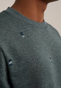 Närbild av en person som bär en grön stickad sweater med små staplade prickmönster i tre färger nära axeln och bröstet.