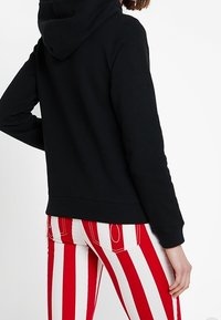 Sudadera con capucha negra de textura suave y capucha, combinada con pantalones rayados en rojo y blanco que muestran un diseño ajustado y costuras visibles.