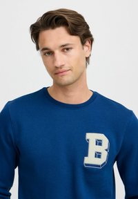 Plava dukserica s okruglim izrezom, sa istaknutim bijelim 3D "B" logom na lijevoj strani prsnog koša. Izrađena od mekane, teksturirane tkanine.