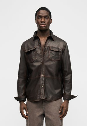 Giovane uomo in piedi, volto rivolto in avanti, che indossa una camicia di pelle marrone scuro con bottoni, tasche sul petto e polsini arrotolati.