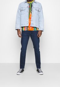 Ljusblå denimjacka över en färgglad mönstrad skjorta, tillsammans med mörkblå jeans och svarta högklackade sneakers med vita sulor.