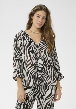 Ung kvinde iført matchende sort-hvid zebra-print bluse med flæser og bukser, accessoriseret med guldringeøreringe og armbånd.