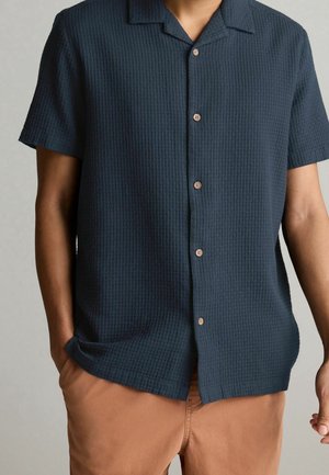 Homme portant une chemise à manches courtes boutonnée bleu foncé texturée avec un pantalon beige, mains détendues, une dans la poche.