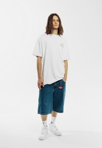 Camiseta blanca de gran tamaño con un pequeño logo, combinada con shorts de mezclilla azules. Zapatillas blancas y calcetines blancos completan el conjunto.