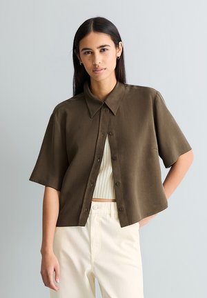 Woman with long dark hair wears an olive green short-sleeve buttoned shirt over a white ribbed crop top and off-white pants, standing against gray.