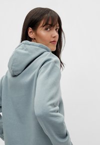 Hanorac cu glugă de culoare albastru deschis, cu o textură moale, croială relaxată și cusături discrete. Părul modelului este lung și drept, acoperindu-i parțial fața.