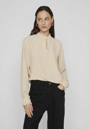 Blusa beige a maniche lunghe con scollatura a keyhole e rifiniture volanti. Abbinata a pantaloni neri a vita alta. Tessuto morbido, vestibilità comoda.