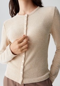 Beige Rippenstrick-Cardigan mit langen Ärmeln, rundem Ausschnitt und Knopfverschluss. Auffällige Textur und weicher Stoff mit dezentem Streifenmuster.