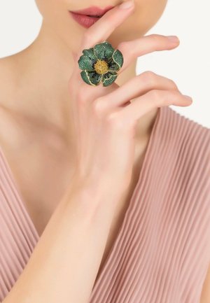 Green and gold flower ring with textured petals, sparkling accents, and a golden center, worn on a hand against a light background.