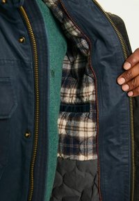 Marineblaue Jacke mit kariertem Futter, Reißverschluss, gesteppter grauer Innenstoff und goldfarbene Beschläge. Darunter ein grüner Pullover sichtbar.