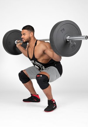 Muscular man wearing knee pads squats with a heavy barbell across his upper back in a white studio setting.