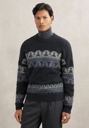 Homme portant un pull foncé et duveteux avec des motifs géométriques bleu-gris sur un col roulé gris, associé à un pantalon gris foncé.