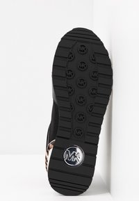 Semelle de chaussure de sport noire avec des motifs de bande de roulement comportant des logos circulaires "MK" et un emblème "MK" argenté près du talon.