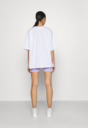 Nainen seisoo selin, yllään ylisuurikokoinen valkoinen T-paita, lila pyöräilyshortsit, valkoiset lenkkarit ja renkaanmuotoiset korvakorut, taustana yksivärinen seinä.