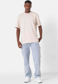 Lys beige bomulds t-shirt med korte ærmer og diskret hvid tekst, sammen med lyseblå denim cargo bukser og hvide sneakers.