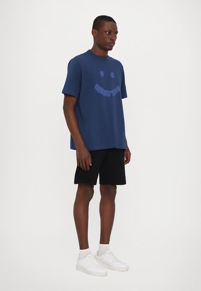 T-shirt en coton marine avec un col rond et un design graphique de smiley. Portée avec un short noir et des baskets blanches. Look simple et décontracté.