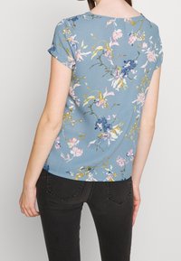 Blusa de manga corta floral hecha de tela azul clara, con flores en tonos rosa, blanco y azul oscuro, y detalles en verde; textura suave.