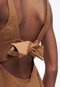 Gros plan d'une personne portant un vêtement marron sans manches avec un nœud en tissu noué dans le dos ouvert.