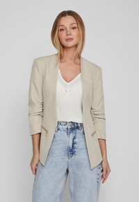 Sacou bej cu un design structurat, două buzunare cu fermoar și mâneci trei sferturi, purtat peste un top alb și blugi cu talie înaltă, decolorati.