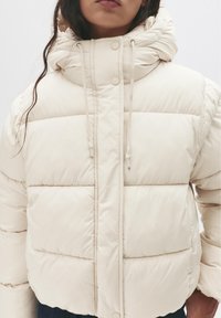 Kobieta ubrana w kremową pikowaną kurtkę z wysokim kołnierzem i troczkami, widoczna od ramion do bioder.