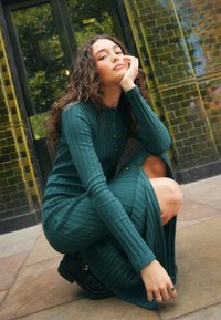 Vestito verde a costine lungo fino al ginocchio con maniche lunghe, caratterizzato da bottoni dorati e uno spacco laterale. Indossato con stivaletti neri alla caviglia.