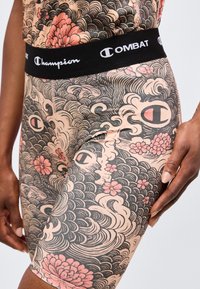 Gros plan sur une personne portant un short Champion Combat à motifs floraux et ondulés, avec une ceinture noire et une main reposant sur la cuisse.