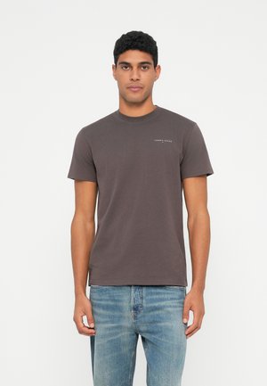 Mann in einem enganliegenden, dunkelgrauen Tommy Jeans T-Shirt und hellblauen Jeans, im Vordergrund eines schlichten weißen Hintergrunds.