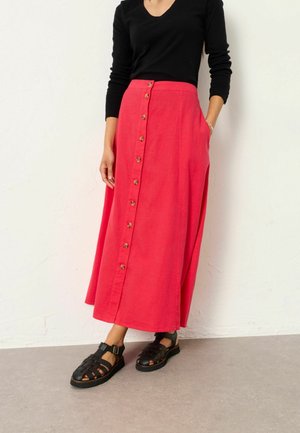 Woman wearing a black long-sleeve top, red button-front midi skirt, and black buckle sandals, standing against a white textured wall.