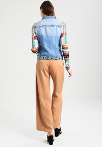 Nainen, jolla on yllään ilman hihnoja vaaleansininen farkkaliivi värikkään kuvioidun pitkähihaisen paidan päällä, vaaleat leveälahkeiset housut ja mustat korkokengät, kääntyy poispäin.