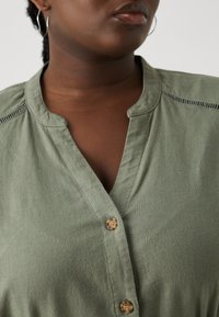 Zielona oliwkowa koszula zapinana na guziki, wykonana z teksturowanej tkaniny, z dekoltem w kształcie litery V, dekoracyjnymi stębnowaniami na ramionach i okrągłymi guzikami.