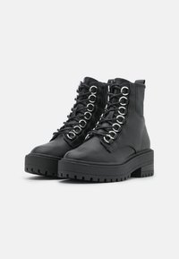 Botas de tobillo de cuero negro con suelas gruesas tipo lug, puntas redondas, múltiples ojales plateados y cordones negros sobre un fondo blanco liso.