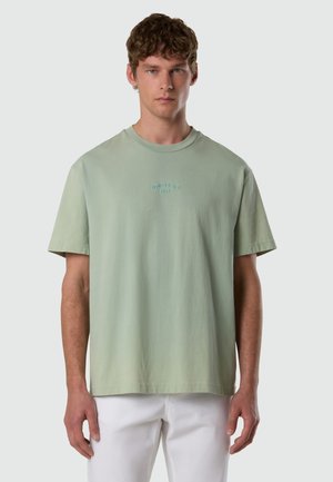 Uomo che indossa una T-shirt verde chiaro con la scritta "North Sails 1957" sul petto e pantaloni bianchi, in piedi davanti a uno sfondo neutro.