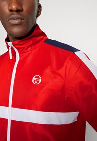 Chaqueta roja de pista con un cuello alto, cremallera blanca y una franja blanca horizontal en el pecho. Detalles en azul marino en los hombros. Logo visible.