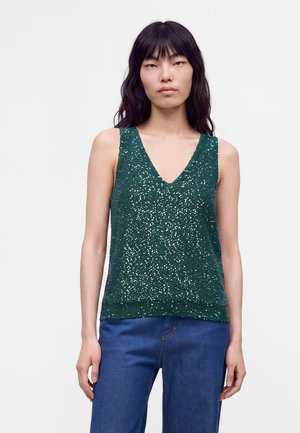Jeune femme aux longs cheveux noirs portant un haut vert à sequins sans manches à col en V et un jean bleu sur un fond clair uni.