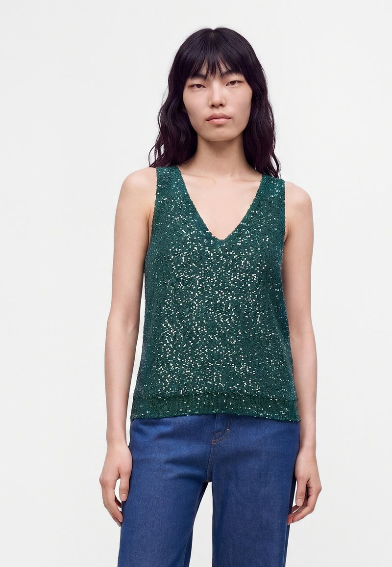 Jeune femme aux longs cheveux noirs portant un haut vert à sequins sans manches à col en V et un jean bleu sur un fond clair uni.
